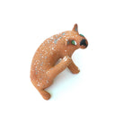 A small sculpted dog figurine in a warm tan-brown glaze with white speckles, a green eye, and a black nose, scratching behind its ear, photographed on a plain white background.
