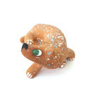 A small sculpted dog figurine in a warm tan-brown glaze with white speckles, a green eye, and a black nose, scratching behind its ear, photographed on a plain white background.