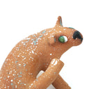 A small sculpted dog figurine in a warm tan-brown glaze with white speckles, a green eye, and a black nose, scratching behind its ear, photographed on a plain white background.