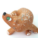 A small sculpted dog figurine in a warm tan-brown glaze with white speckles, a green eye, and a black nose, scratching behind its ear, photographed on a plain white background.