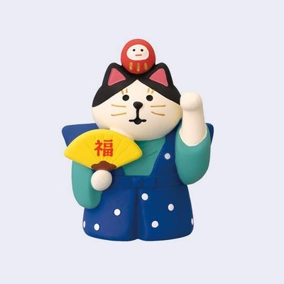 Cat figurine in a blue outfit with a red daruma atop its head and yellow fan on a light purple background