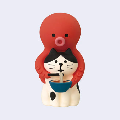 Cute cat figure with a red octopus sitting atop its head, holding chopsticks and feeding the cat a bowl of ramen.