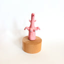 Figure of a pink bird like creature, with tiny wings and boobs with blue nipples. It sits on a wooden circular base.
