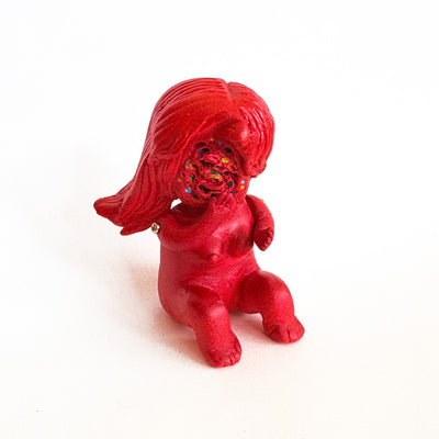 Small figure, all red, of a woman sitting nude on the ground. She has no face, just a swirl where her face would be with dots of colors.