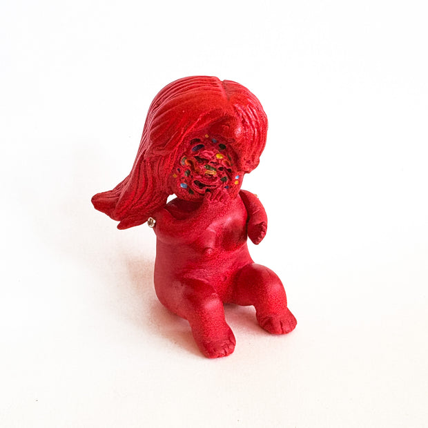 Small figure, all red, of a woman sitting nude on the ground. She has no face, just a swirl where her face would be with dots of colors.
