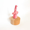 Figure of a pink bird like creature, with tiny wings and boobs with blue nipples. It sits on a wooden circular base.