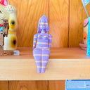 Figure of a woman, all purple with yellow stripes, sitting on a shelf.