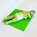 Clay sculpture of an old woman with green skin wearing a one piece bathing suit and laying on her stomach, as if sun bathing. She rests on a green tile.