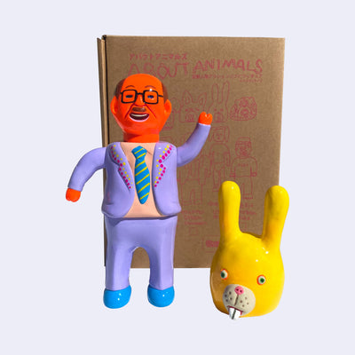 Neon orange vinyl figure of a business man, wearing a lilac colored suit with a peach colored shirt and a blue and yellow striped tie. The lapel of his jacket is decorated with pink and yellow dots. At its feet is a yellow rabbit mask, which can cover the man's whole head.