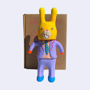Neon orange vinyl figure of a business man, wearing a lilac colored suit with a peach colored shirt and a blue and yellow striped tie. The lapel of his jacket is decorated with pink and yellow dots. On his head is a yellow rabbit mask, covering the entirety of his head and face.