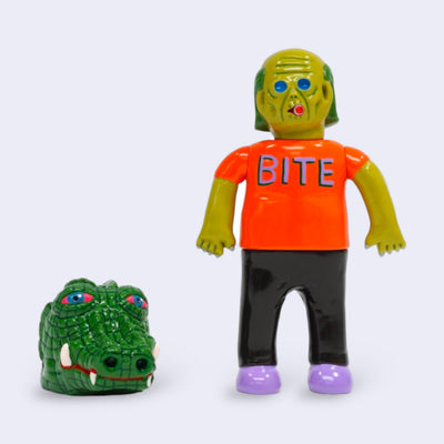 Vinyl figure of a green man, with sunken blue eyes and wrinkled skin akin to a zombie. He smokes a cigar and wears a shirt that reads "bite." Nearby is a large crocodile head, with red glazed over eyes.