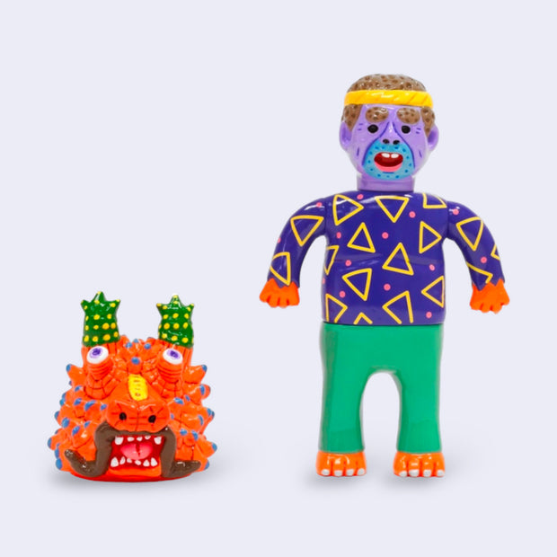 Vinyl figure of a purple man with a braided headband and blue 5 o clock shadow. He wears a bright purple shirt with triangles and green pants. Near him is a dragon head, orange with wild eyes and many horns on its head and face.