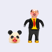 Vinyl doll of a business man, with a black suit, yellow shirt and red tie. He wears golden shoes and has bear paws for hands. Next to him is a large panda head with its mouth open.