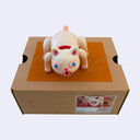 White vinyl figure of a cat, with large blue eyes, an open mouth and textured fur. It stands atop of a cardboard box.