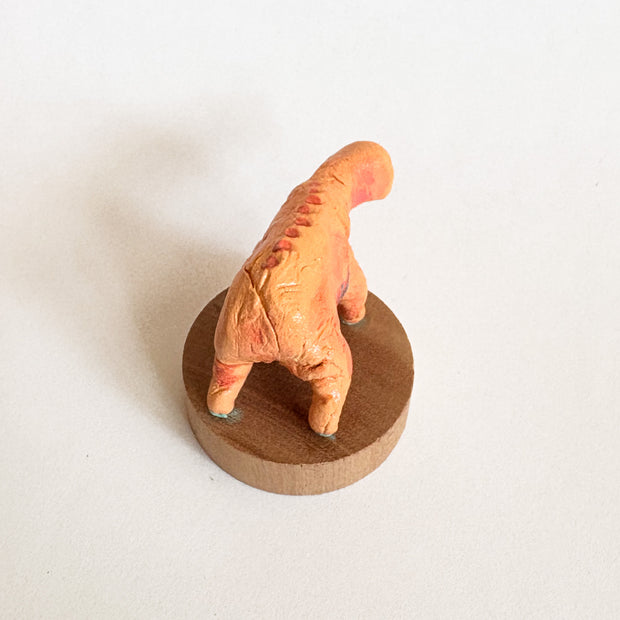 Small orange creature figure with four legs and no face, only an open mouth with many teeth. It stands on a wooden base against a white background