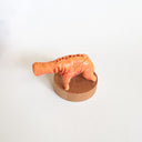Small orange creature figure with four legs and no face, only an open mouth with many teeth. It stands on a wooden base against a white background