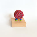 Colorful figurine with a red textured circular spore shape for its body and tiny legs, on a wooden block against a white background