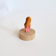 Tiny nude figurine of a person with a pinkish body and long, light-brown hair on a wooden base against a white background