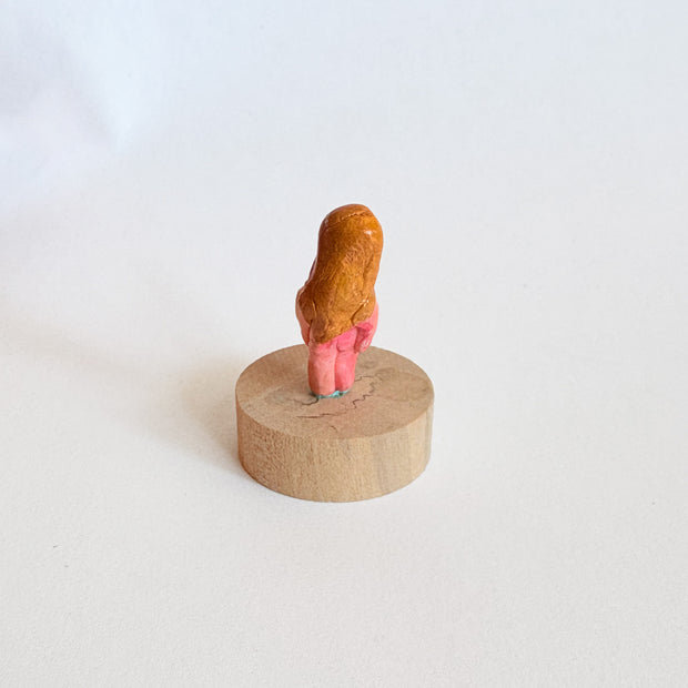 Tiny nude figurine of a person with a pinkish body and long, light-brown hair on a wooden base against a white background