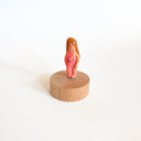 Tiny nude figurine of a person with a pinkish body and long, light-brown hair on a wooden base against a white background