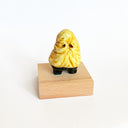 Small yellow sculpture of a ghost-shaped figure with only eyes and tiny legs, wearing black pants and standing on a wooden base.