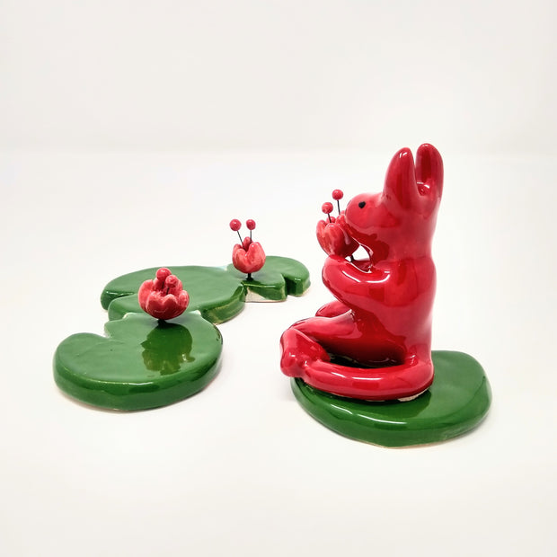 Ceramic sculptures, a cute red devil sits atop of a green lily pad and holds a flower in its hands. Across, is a set of 3 lily pads with flowers blooming out from them.