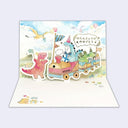 A 3D pop-up greeting card featuring a whimsical dinosaur-themed train scene. The illustration shows a pink dinosaur standing next to a pastel-colored train carrying blue, green, and yellow dinosaurs under a light blue sky with clouds and balloons.