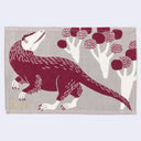 Beige colored rectangle fabric placemat with a burgundy illustration of a feathered raptor-like dinosaur, standing on its hind legs with short arms. It looks back over its shoulder at a series of rounded trees.