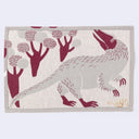 Beige colored rectangle fabric placemat with a burgundy illustration of a feathered raptor-like dinosaur, standing on its hind legs with short arms. It looks back over its shoulder at a series of rounded trees. Reverse side shows inverted colors.