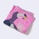 Bright pink square shaped handkerchief featuring imagery of a Dodo bird on the front, eating a green apple in its beak. Next to it is a beige tree with navy blue rounded bunches of leaves. Close up to show embroidered detailing.