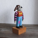 Painted whittled wooden sculpture of a cartoon style dog, standing up like a human and wearing a striped shirt and jeans. It has long, floppy ears and is half brown and half white.