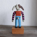 Painted whittled wooden sculpture of a cartoon style dog, standing up like a human and wearing a striped shirt and jeans. It has long, floppy ears and is half brown and half white.