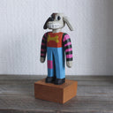 Painted whittled wooden sculpture of a cartoon style dog, standing up like a human and wearing a striped shirt and jeans. It has long, floppy ears and is half brown and half white.
