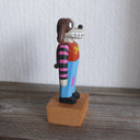 Painted whittled wooden sculpture of a cartoon style dog, standing up like a human and wearing a striped shirt and jeans. It has long, floppy ears and is half brown and half white.