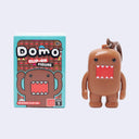 A Domo clip-on figure shown next to its retail box on a light gray background. The figure is brown with a red mouth showing white teeth and a small brown hair tuft, attached to a metal key ring.