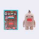 A Domo clip-on flocked figure shown next to its retail box on a light gray background. The box features the 'Domo Clip-On' branding and an illustration of the character, while the plush itself is a pale pinkish-beige color with a red mouth and black eyes.
