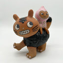 Ceramic sculpture of a brown cat-like creature with a toothy grin and looking at the top of its own back, where a character sits and wears a pink body suit.