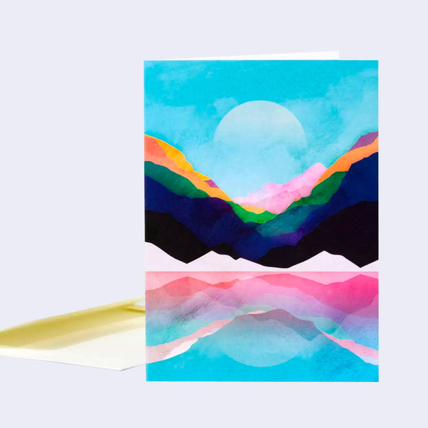 Greeting card with a colorful illustration of simple colored mountains reflecting onto a blue lake. The moon is very light in the sky, appearing in mid day.
