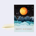 Greeting card featuring an illustration of a large orange moon in a dark starry sky over blue mountains. The mountain range  furthest back is realistic in style and the ones in front are cut out of paper. Text reads "happy birthday"