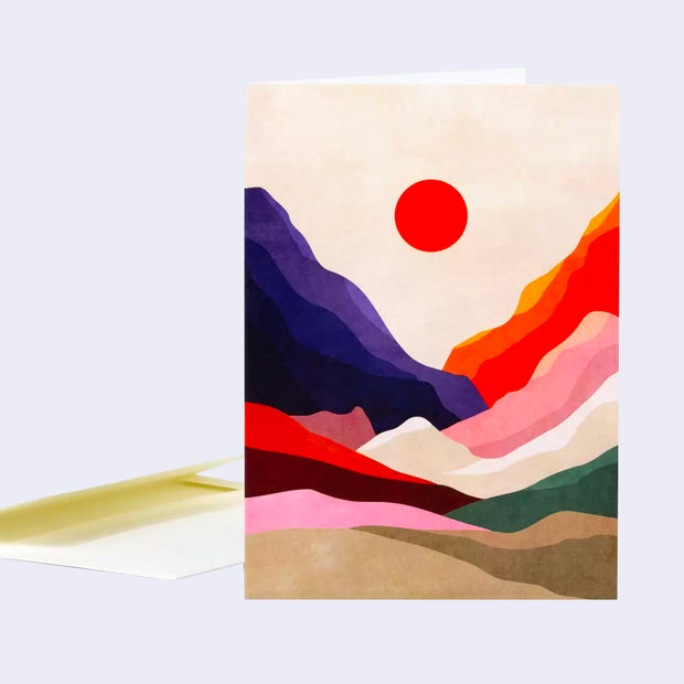 Notecard with a simplistic mountain scene, with various colorful layers of valleys and a large red sun in the middle against a cream sky. Envelope sits nearby.
