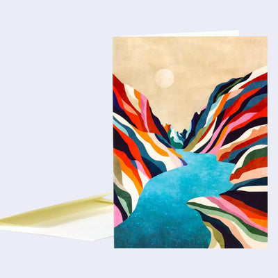 Greeting card featuring an illustration of a large lake, flowing in between 2 mountains that are striped with many different colorful but muted colorways. The sky is tan with a small moon or sun hanging overhead.