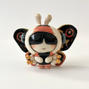 Ceramic sculpture of a small character with a large head wearing a Mothra costume, with a hood hat and a large set of wings behind it matching Mothra's colorways.