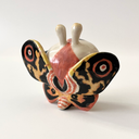 Ceramic sculpture of a small character with a large head wearing a Mothra costume, with a hood hat and a large set of wings behind it matching Mothra's colorways.