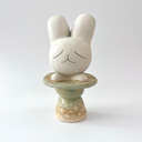 Ceramic sculpture of a platform, akin to the bottom of a candle holder, with a white bunny character popping out of the top.