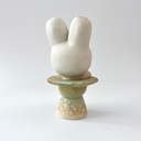 Ceramic sculpture of a platform, akin to the bottom of a candle holder, with a white bunny character popping out of the top.