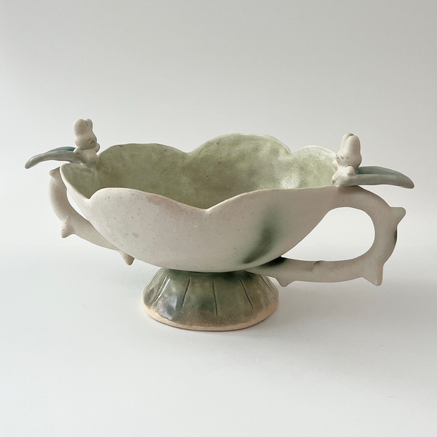 Ceramic vessel, a wide teacup with 2 handles. The bowl of the cup is shaped like a flower and the handles have thorns on them. Atop each handle is a small white bunny character.