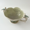 Ceramic vessel, a wide teacup with 2 handles. The bowl of the cup is shaped like a flower and the handles have thorns on them. Atop each handle is a small white bunny character.