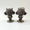 2 dark brown ceramic sculptures of a character. One with a very large head and a small body. It has earring cuffs, a halo, and holds a small white flower. The other has horns and also holds a flower.