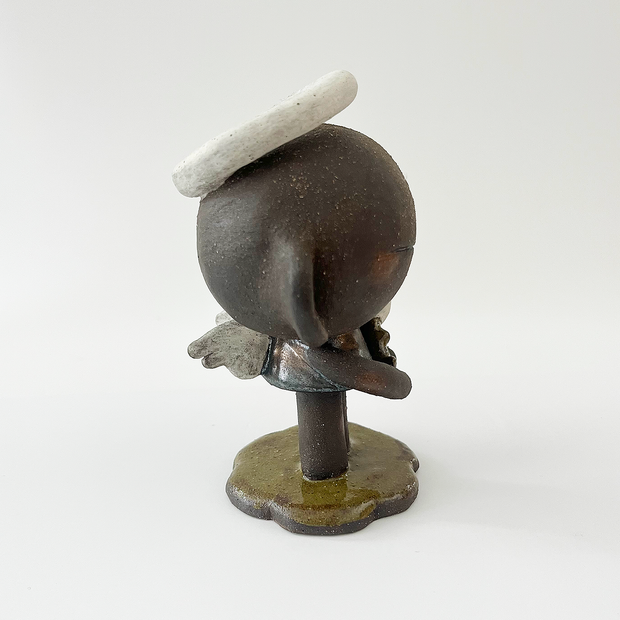 Dark brown ceramic sculpture of a character with a very large head and a small body. It has earring cuffs, a halo, and holds a small white flower.