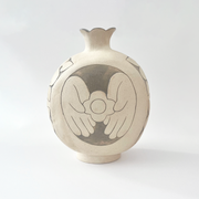 Cream colored ceramic vase with a very round body and a small fluted top, almost resembling a pomegranate in shape. On the back is a design of a person's hands holding a flower.